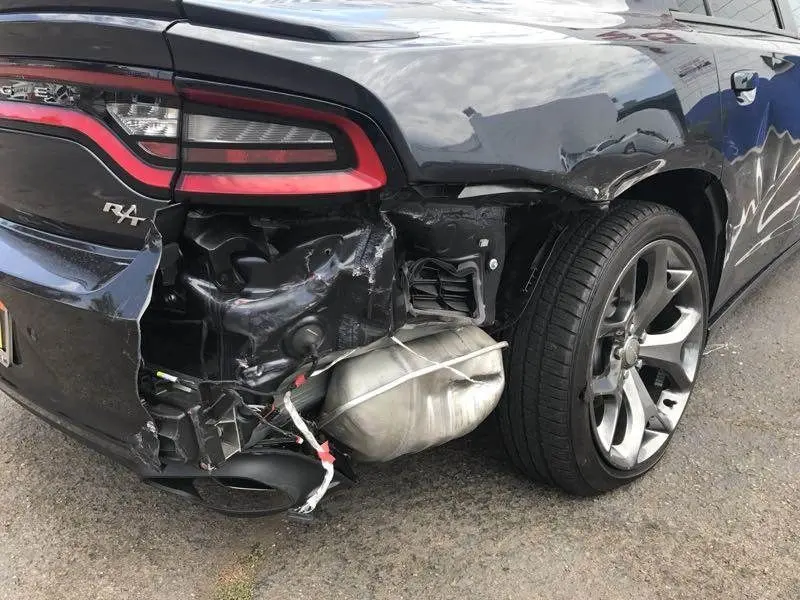 The rear quarter panel of a dark Dodge Charger R/T torn open after a collision, with the bumper cover hanging away, the exhaust and rear suspension exposed, and the tail light assembly pushed out of line