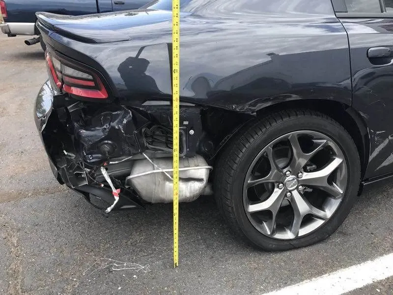 A tape measure held vertically against the torn rear quarter of the Dodge Charger to record how far the panel has moved, with the crushed bumper and exposed exhaust behind it