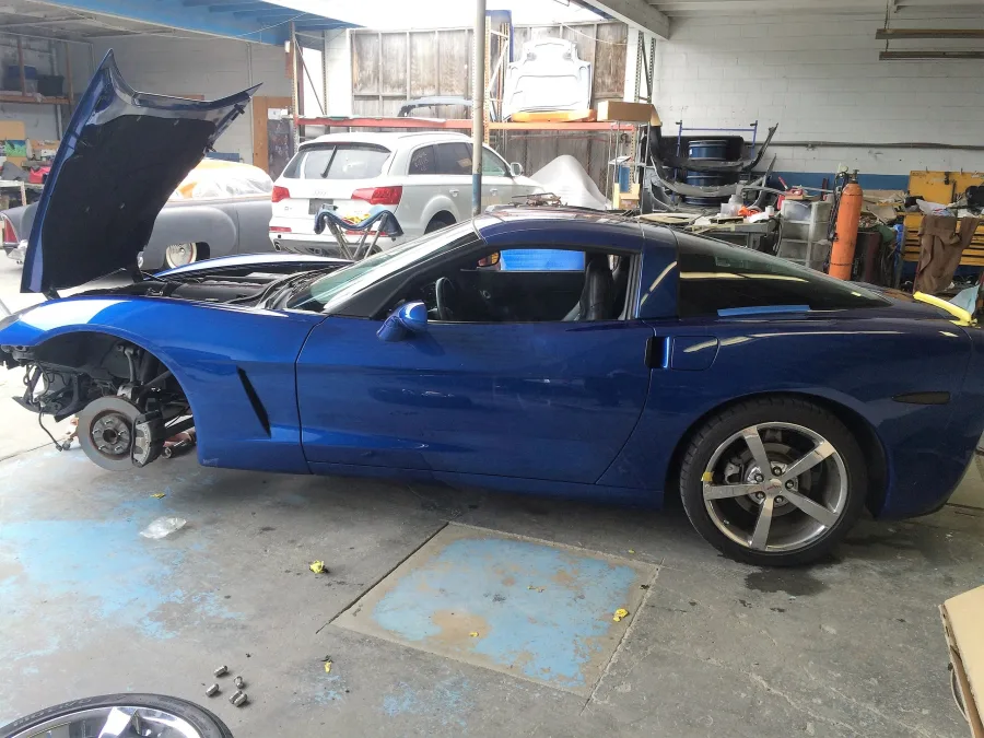 Inside the Elite Auto Collision bay, the blue Corvette up on its wheels with the hood open and the front end disassembled, parts racks and equipment lining the wall behind it