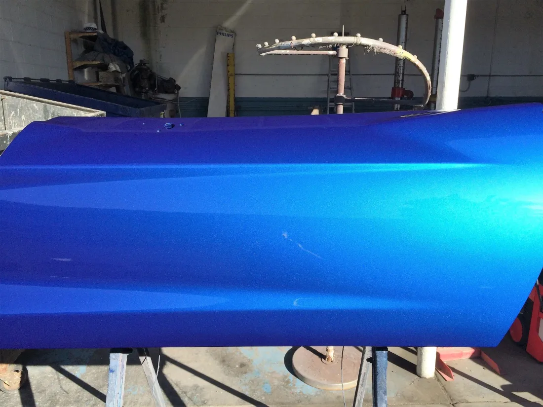 A metallic blue door panel just out of the booth, its wet finish catching the light along the whole length of the panel